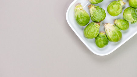 A close-up image of fresh green brussels sprouts in a white container on a gray background. They are round and green with a slight purple tint, neatly arranged and well-lit.の写真素材