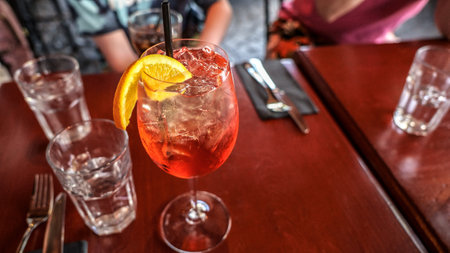 This close-up image features a single sparkling wine glass, embellished with a bright orange slice. It is placed on a dark red table, accompanied by two glasses of water, a knife, and a fork.の写真素材