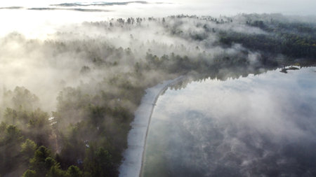 The image shows a beautiful lakeside campsite in the morning. The campsite is surrounded by lush green trees and the lake is shrouded in a thin layer of fog, creating a peaceful and serene atmosphere.の写真素材