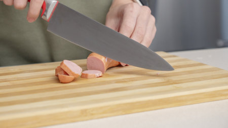 Close-up of a woman cutting sausage on a wooden cutting board in a kitchen setting with different ingredientsの写真素材
