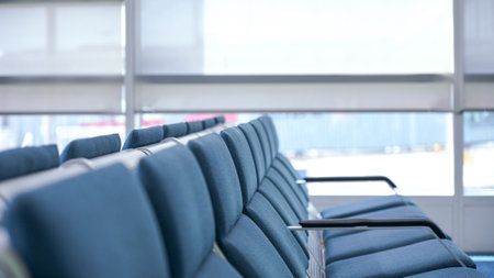 This is a photo of a row of empty blue airport seats with a large glass window in the background. The photo is taken from the perspective of a passenger sitting in one of the seats.の写真素材