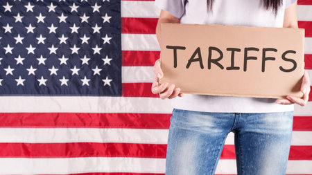 A young woman stands before an American flag, holding a sign that says tariffs in black marker. She wears a white shirt and blue jeans. The photo is from a low angle, concealing her face.の写真素材
