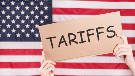A cardboard sign with the word Tariffs written on it is held up in front of an American flag. The sign is held by a pair of hands and the background is out of focus.の写真素材