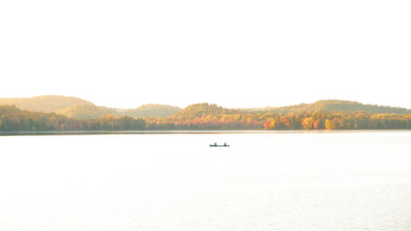 The image shows a small boat on a calm lake with a mountain range in the background. The boat is rowed by two people and the sun is rising or setting, casting a golden glow over the scene.の写真素材