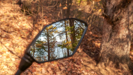 Looking through the rearview mirror of a motorcycle, you see a beautiful view of a lake surrounded by trees in the distance. The dirt road through the forest is covered with fallen leaves.の写真素材
