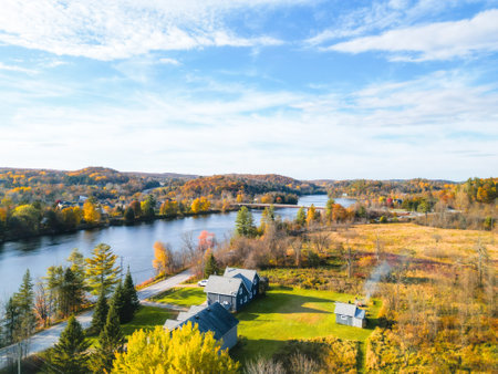 A scenic rural landscape showcases a flowing river, vibrant autumn trees, and a cozy house by the water. The clear blue sky with white clouds enhances the peaceful atmosphere.の写真素材