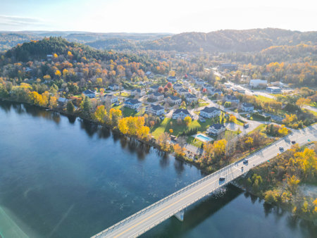 A beautiful small town filled with charming houses, vibrant trees, and a serene river flowing through it. The sky is a brilliant blue adorned with fluffy white clouds, and the sun shines brightly.の写真素材