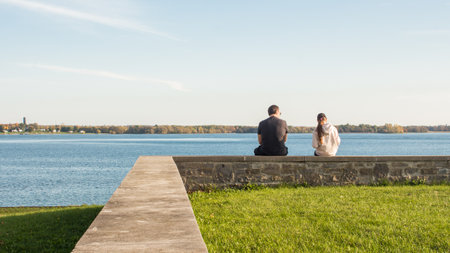 A young couple is sitting on a stone wall overlooking a tranquil lake. They are gazing out at the stunning view and enjoying the peace and quiet, surrounded by nature s beauty and serenity.の写真素材
