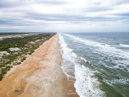 The beach is wide and sandy with a gentle slope into the ocean. The water is clear and blue-green. The sky is blue with scattered clouds. There are dunes covered with sea oats behind the beach.の写真素材