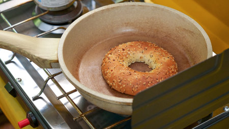 The bread is a bagel with sesame seeds on top. The ceramic pan is brown and has a wooden handle. The camp stove is silver and has a black grate on top. The bagel is round and has a hole in the middle.の写真素材