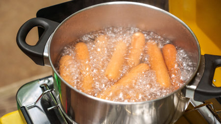The image features a shiny stainless steel pot filled with bubbling boiling water and vibrant orange carrots. The pot sits atop a portable camping gas burner, showcasing a practical cooking setup.の写真素材