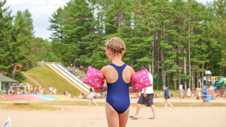 Little girl with pink water wings standing on the sandy beach near the playground on a sunny summer day. The background is blurred, and the girl is wearing a blue one-piece swimsuit.の写真素材