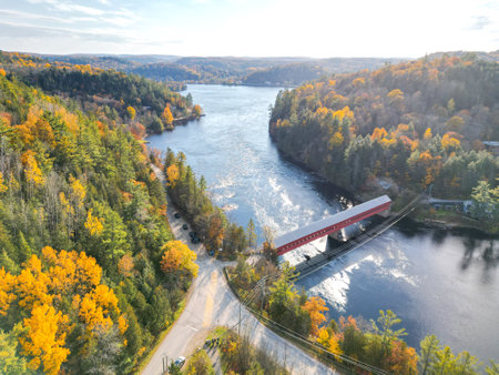 The image shows a small red bridge over a river in a beautiful autumn forest. The trees are in full foliage and the leaves are turning yellow, orange, and red. The water is calm and crystal clear.の写真素材