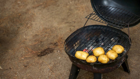 A selection of unwashed potatoes are placed on a charcoal grill, cooking with the lid open. The smoky aroma fills the air as the potatoes sizzle on the grate, showcasing a delicious grilling process.の写真素材