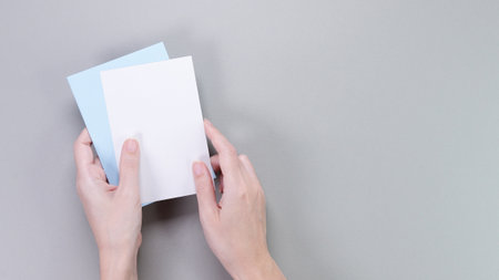 The image showcases a pair of slender female hands elegantly holding two blank note cards slightly apart. The cards are set against a solid gray background, creating a clean and modern aesthetic.の写真素材