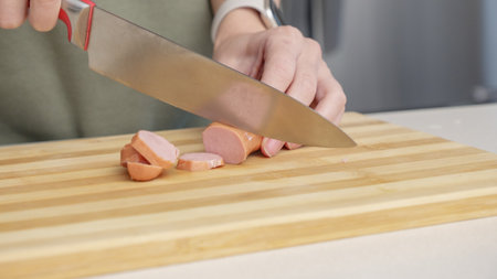 Close-up of a woman cutting sausage on a wooden cutting board in a kitchen setting with different ingredientsの写真素材