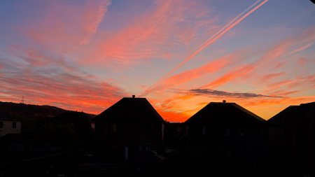 A stunning sunset illuminates the sky with shades of orange and pink over suburban rooftops. Vapor trails create accents across this serene urban landscape, offering a picturesque view.の写真素材