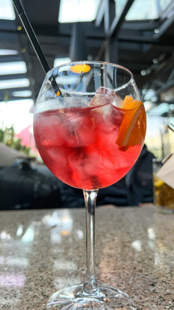 A close-up of a refreshing red cocktail with ice cubes and an orange slice in a wine glass on a bar counter, creating a vibrant summer drink experience. Perfect for menus and promotions.の写真素材