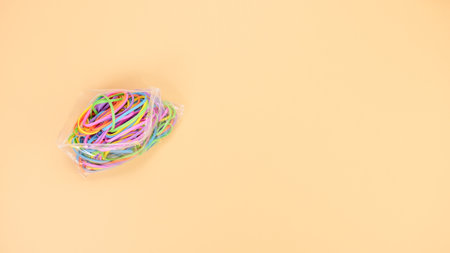 A vibrant assortment of small, thin, circular rubber bands in various bright colors is neatly packed in a transparent plastic bag. The bag lies flat on a smooth, solid peach-colored surface.の写真素材