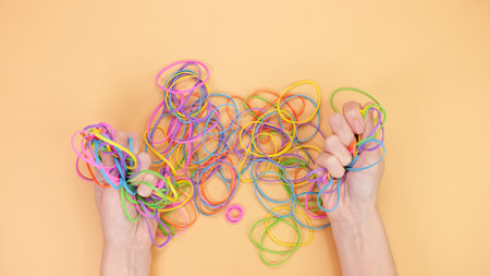 This image showcases a vibrant array of different colored rubber bands intertwined and tangled together on a soft peach-colored background, creating a visually appealing and artistic display.の写真素材