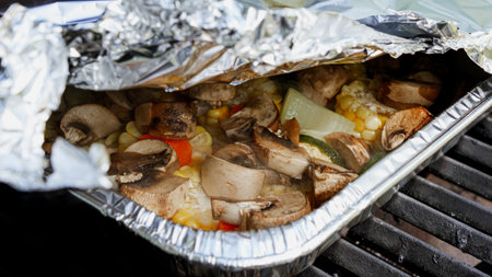 A close-up of an aluminum foil packet with grilled vegetables including mushrooms, zucchini, corn, and peppers on a barbecue grill, showcasing a healthy outdoor summer cooking scene.の写真素材