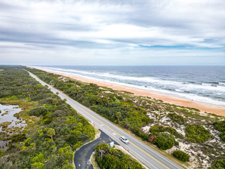 A picturesque coastal highway winds along a pristine beach with soft white sand and calm waves, bordered by vibrant greenery and the expansive ocean, creating a serene and inviting atmosphere.の写真素材