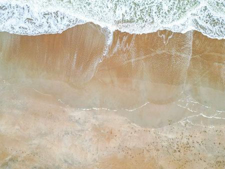 This image features a peaceful sandy beach with light brown sand. Gentle waves lap at the shore, and the clear blue water sparkles under the sun, creating a serene and inviting atmosphere.の写真素材