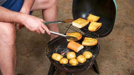 A close-up shot of a charcoal grill with a grate full of potatoes and garlic bread. The potatoes are various sizes and the garlic bread is made with thick slices of bread.の写真素材