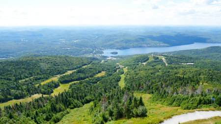 A lush green mountain peak offers a breathtaking view of a tranquil lake and a charming village nestled in the distance. The sky is clear with a few fluffy white clouds drifting peacefully.の写真素材