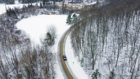 An aerial view captures a car driving along a winding road through a snowy winter landscape. The road cuts through a forest with snow-covered trees and a field, showcasing a serene travel scene.の写真素材