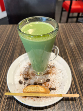 Refreshing green drink presented in a tall glass on a white plate, accompanied by a cookie and coffee beans, creating a cozy cafe atmosphereの写真素材