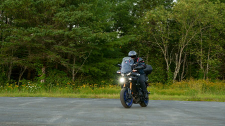 A person confidently sits on a sleek motorcycle in a spacious parking lot. They wear a stylish black jacket and a helmet with a clear visor, while a chainlink fence and lush trees form the backdrop.の写真素材