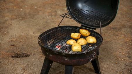 A selection of unwashed potatoes are placed on a charcoal grill, cooking with the lid open. The smoky aroma fills the air as the potatoes sizzle on the grate, showcasing a delicious grilling process.の写真素材