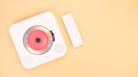 The image features a sleek white portable CD player accompanied by a remote control, set against a soft peach-colored background, with a vibrant red light illuminating the disc compartment.の写真素材