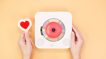 A top-down view of hands forming a heart shape around a modern white CD player on a yellow background. This image conveys themes of love, music, and creativity, perfect for blogs.の写真素材