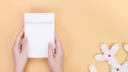 Close up of hands holding a blank white envelope on a beige background with fluffy white Christmas...