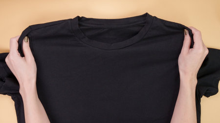 A sleek black t-shirt is showcased against a beige background, with hands holding the shoulders. Perfect for fashion mockups, clothing catalogs, or online retail displays.の写真素材
