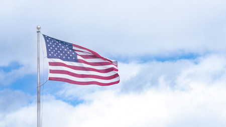 American flag flutters in the wind, set against a bright blue sky with fluffy white clouds,...