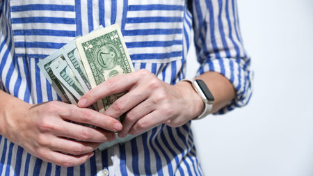 A person in a striped shirt holds a stack of United States dollar bills, symbolizing financial...
