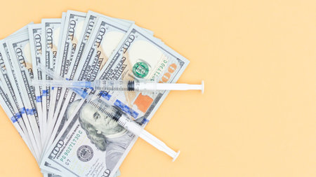 A stack of hundred dollar bills with syringes placed on top, set against a solid color background. Highlights healthcare costs and the financial burden of medical treatment.の写真素材
