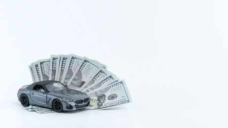 Concept for financial planning to buy a car or saving money for a new automobile. Isolated modern toy car stands beside a stack of 100 dollar bills, isolated on a white solid background.の写真素材