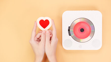 Top view of hands holding a heart-shaped object next to a compact disc player on a yellow background. This image symbolizes love for music and makes a great gift idea.の写真素材