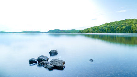Tranquil lake scene featuring smooth water reflecting green hills and scattered rocks, evoking a...