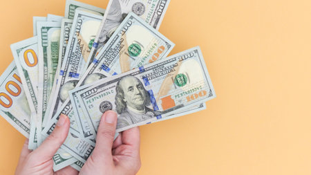 A hand holding a stack of hundred dollar bills against a peach background. Symbolizes wealth,...