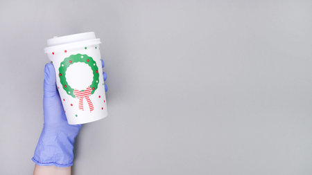 A gloved hand presents a decorative coffee cup featuring a holiday wreath and ribbon, set against a...