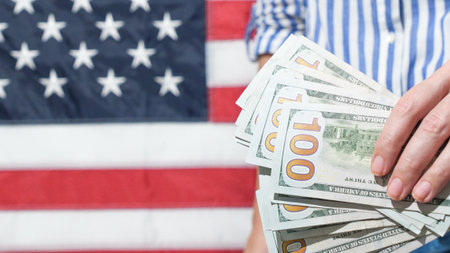 A hand holds a stack of hundred dollar bills in front of a blurred American flag, representing wealth, finance, and the American dream. Perfect for economic themes and financial success.の写真素材