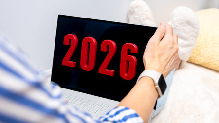 A person with light skin holds a laptop with digital display showing the year 2026. The setting is cozy, with soft textures and warm colorsの写真素材