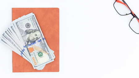 A stack of one hundred dollar bills sits on an orange notepad beside red framed glasses on a white...