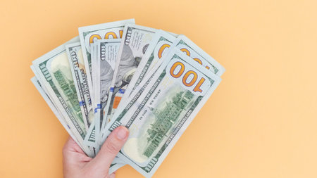 Close up of a hand holding a stack of US hundred dollar bills against a solid yellow background....