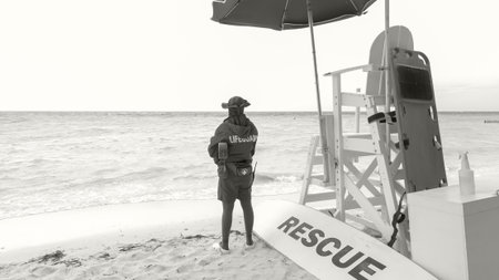 Lifeguard in uniform stands on sandy beach near rescue equipment, overlooking calm ocean waves under an umbrella, ensuring safety for beachgoers.の写真素材